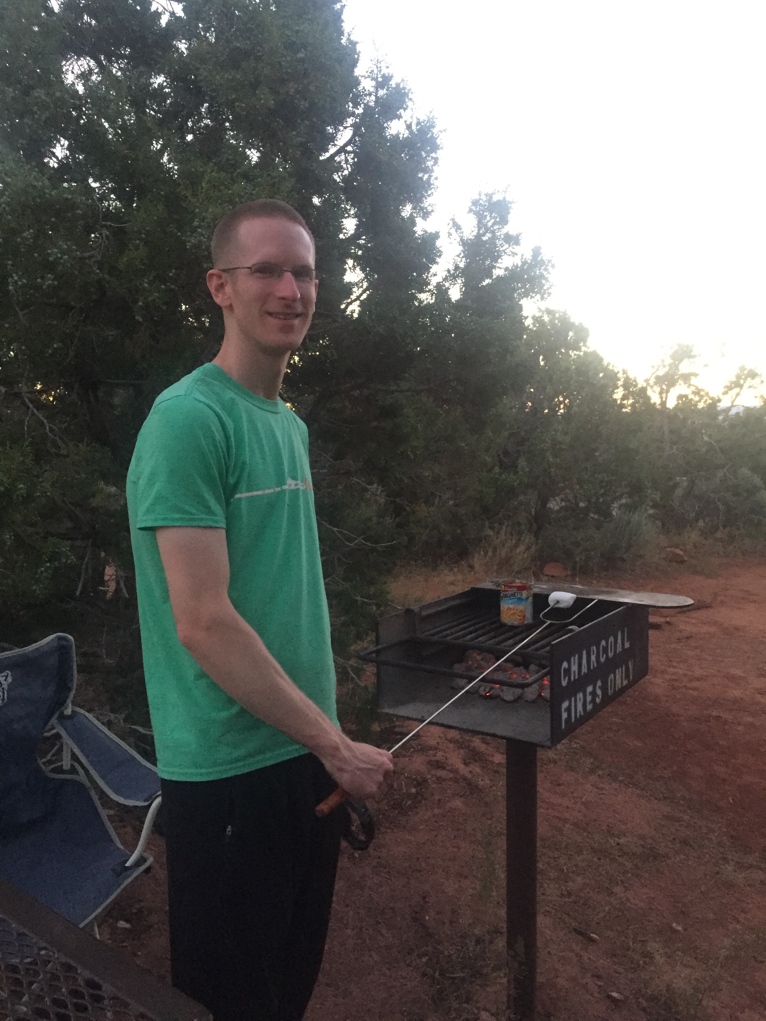 Camping at Colorado National Monument Saddlehorn Campground. A white man stands in front of a basic charcoal grill, holding a metal skewer with a marshmallow on the end.