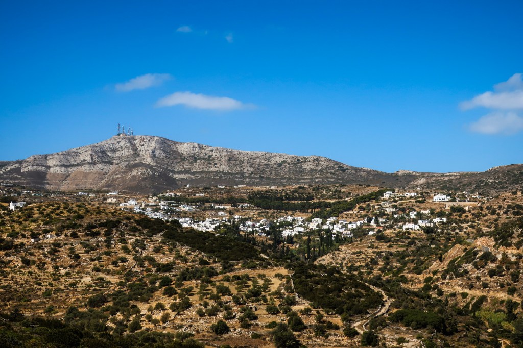 Scrubby mountains with the white buildings of Lefkes visible in a small valley. A mountain peak is on the horizon with several radio towers poking out into the clear blue sky.