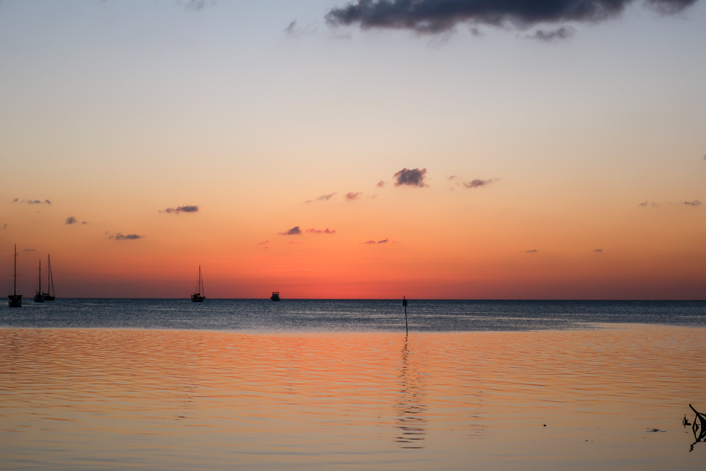 the sky is lit up orange at sunset in caye caulker belize. sailboats are visible on the horizon and the sunset reflects off the water.