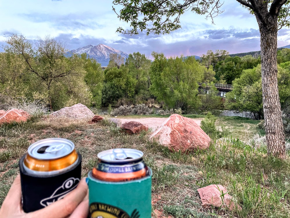 views of roaring fork river and mt sopris with two people cheersing beers in the foreground