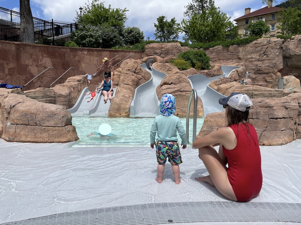 a toddler faces away from the camera toward a set of three water slides while their parent sits next to them