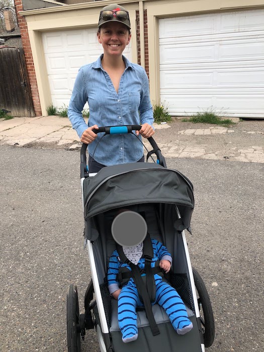 A white woman stands behind the Thule Urban Glide running stroller with a baby strapped into it, both facing the camera.